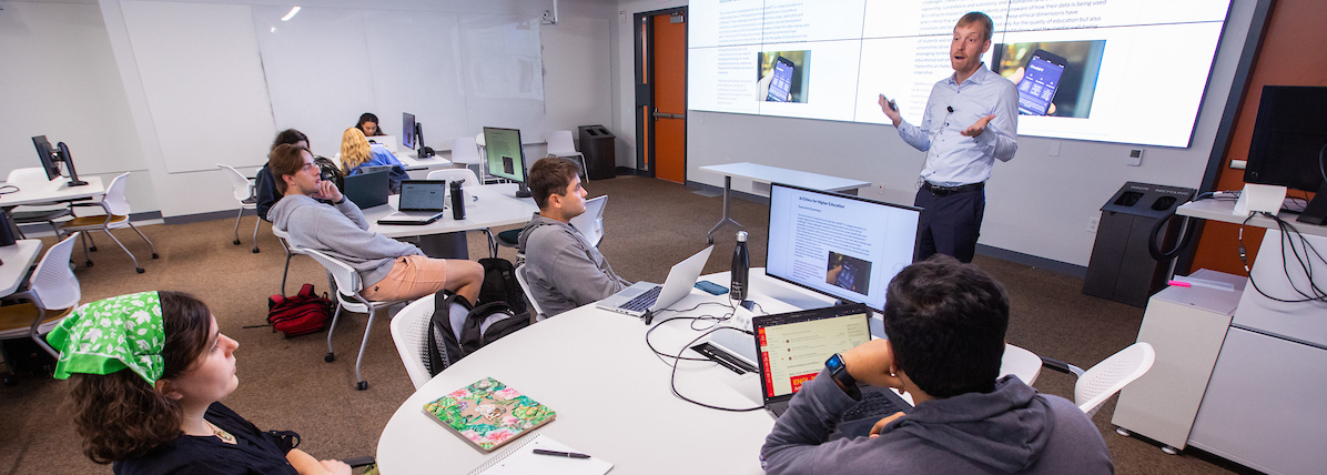 Professor gestures as he speaks to students in front of a large monitor with AI Ethics information