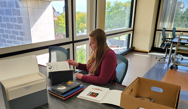 Graphic: Rectangle color image of SCUA reading room with Rebecca Wells, who has long brown hair. She is wearing a long sleeved red shirt and is sitting at desk with book open looking at a page, surrounded by records boxes and other books.